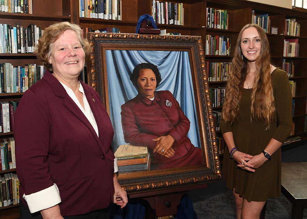 Two women with a portrait of a woman in the libary.
