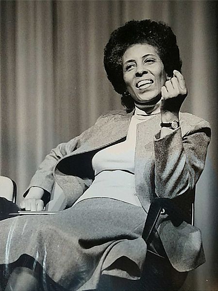Black and white photo of a woman laughing, wearing a blazer, with one hand up