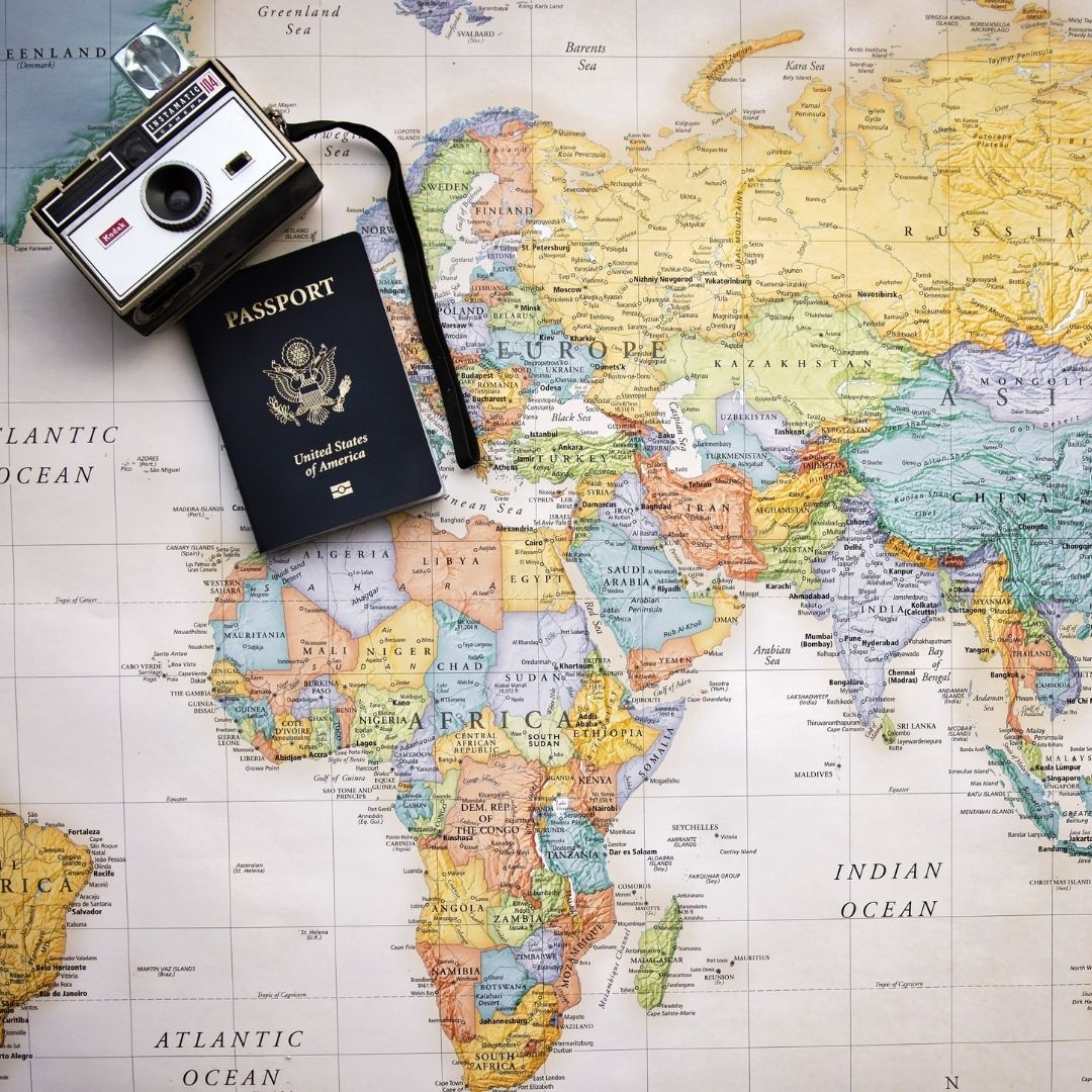 A colorful map of the world as a background, with a United States passport and a silver digital camera laying on top in the upper left of the screen.