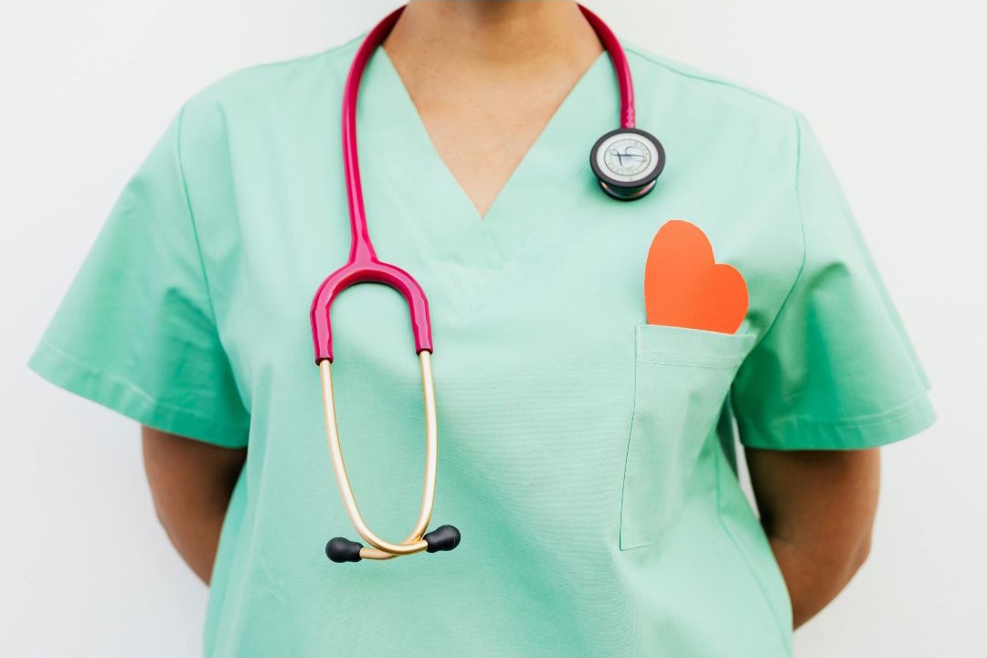 Image of a healthcare professional wearing mint green scrubs with a pink corded stethascope over chest and orange heart in scrub chest pocket